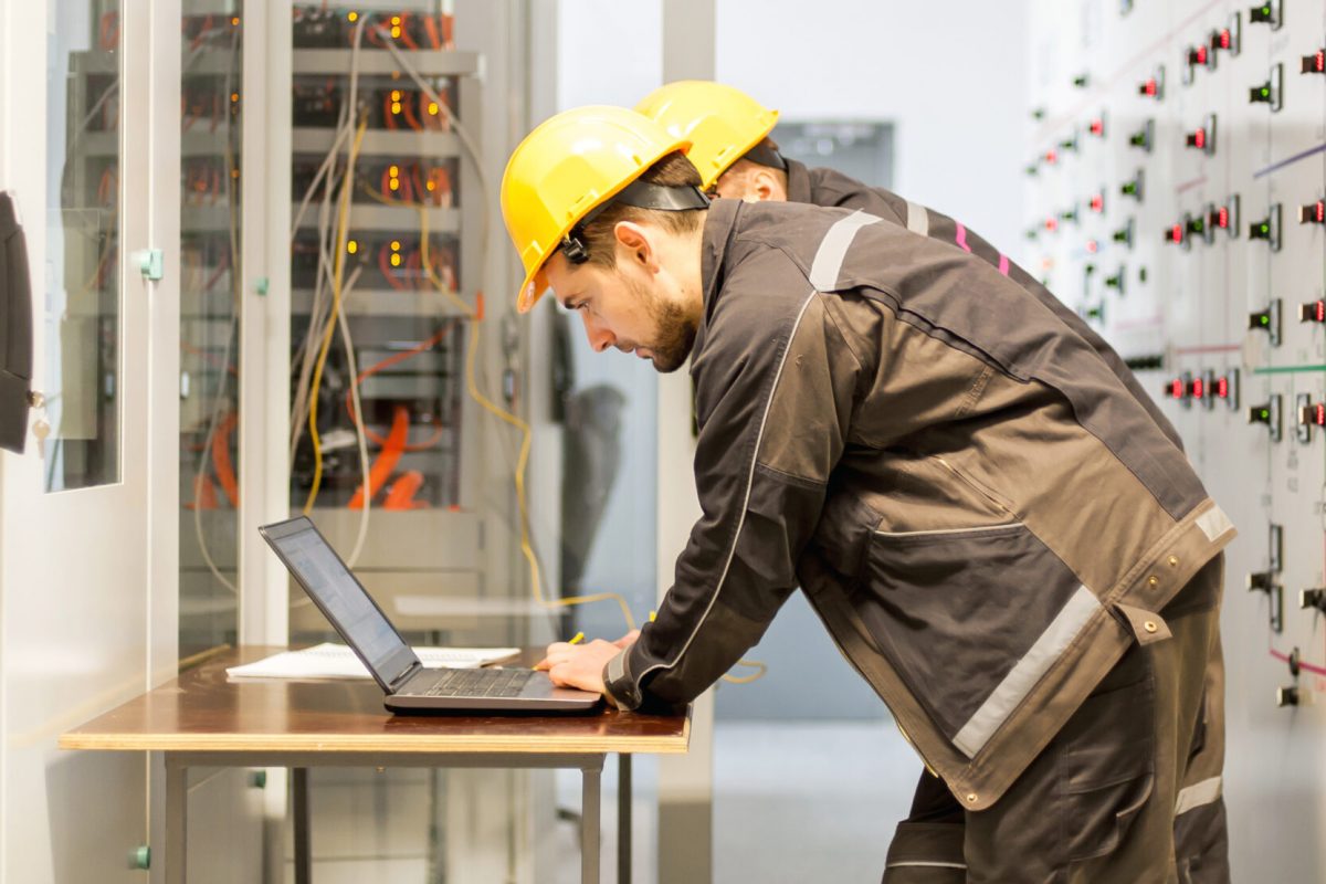 Two maintenance engineers inspect relay protection system with laptop computer. Bay control unit. Medium voltage switchgear